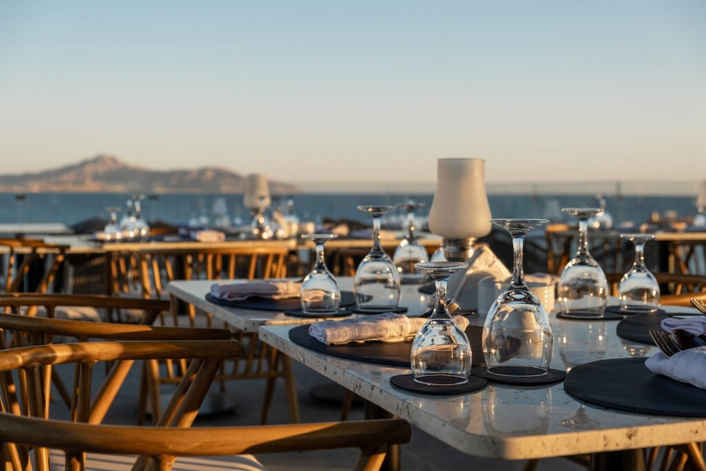 A restaurant terrace with tables set up and glasses of wine, overlooking the sea at sunset